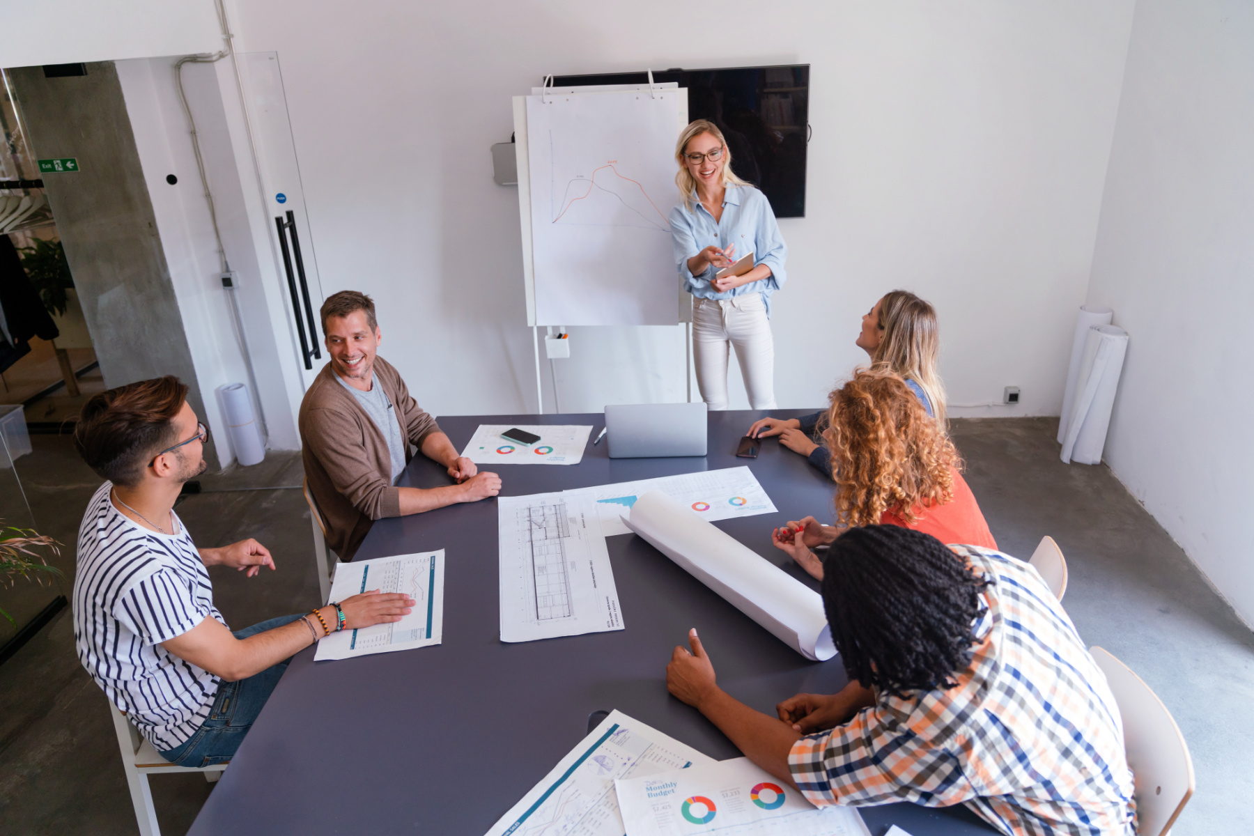 A woman stands by a flip chart with a graph, presenting the planning stage of the AI automation process to five colleagues seated around a table with charts and documents in a modern office.
