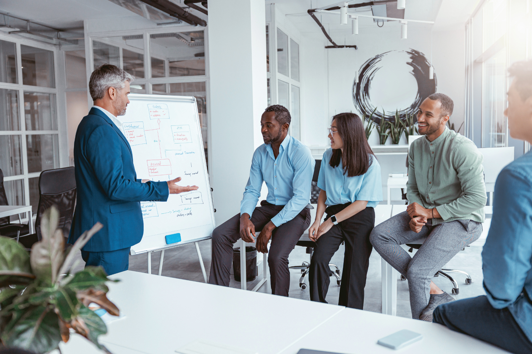 A man in a suit presents to four colleagues seated near a whiteboard with diagrams, discussing intelligent workflow solutions in a modern office setting.