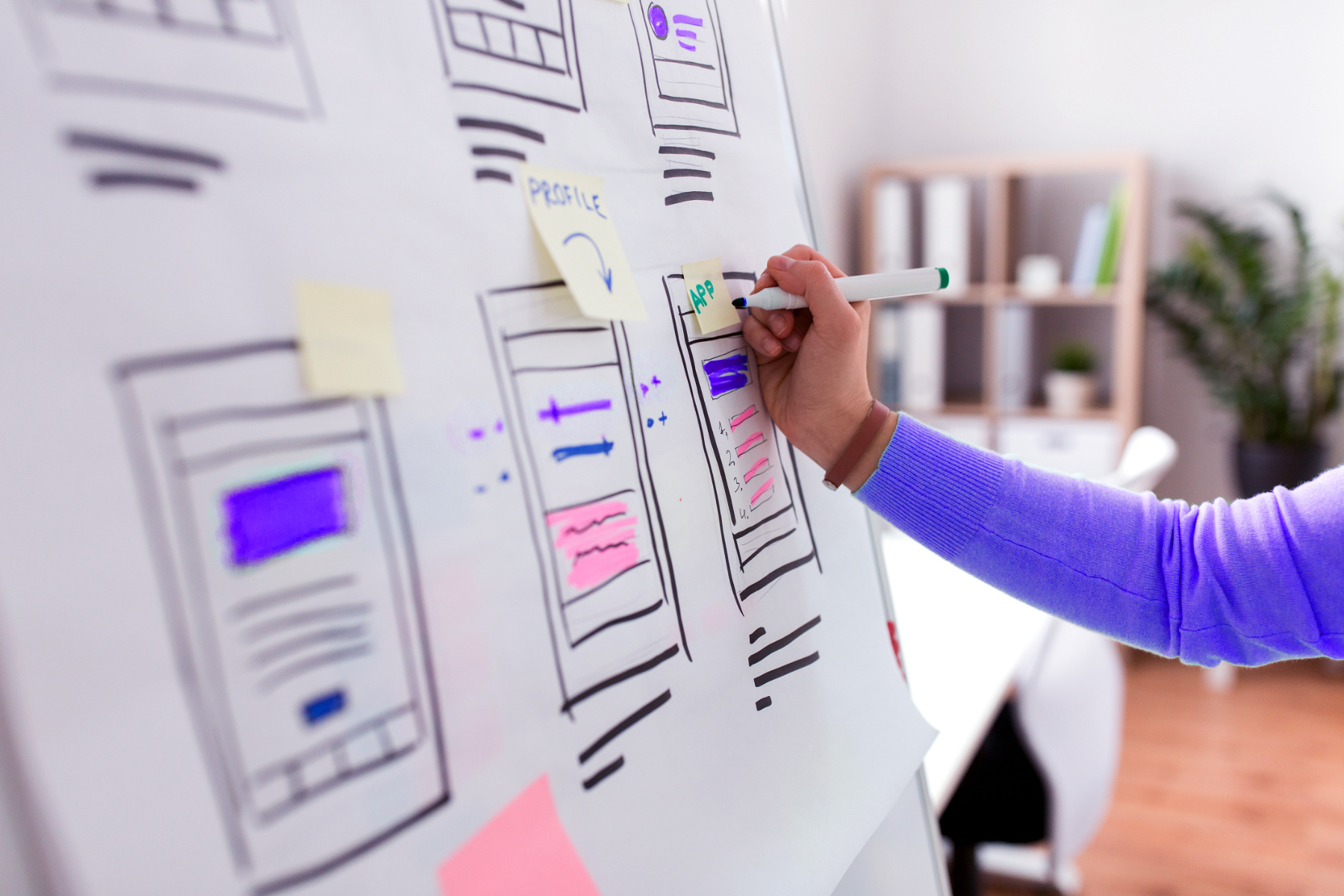 A person sketches wireframes with a marker on a large paper, planning website layouts using sticky notes and diagrams—a glimpse into clean process and UX/UI design in a bright office setting.